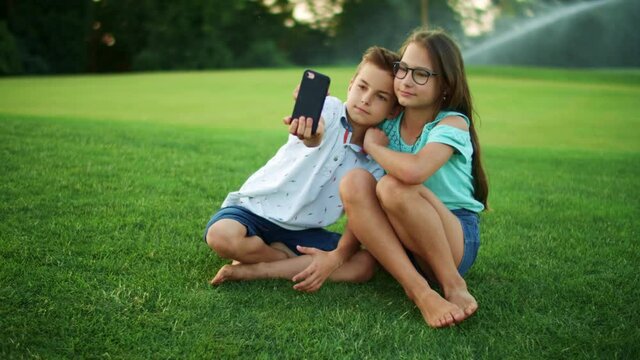 Sister hugging brother in park. Smiling boy taking selfie on cellphone with girl