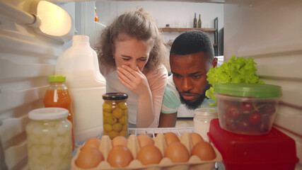 View from inside fridge of young multiracial couple feeling bad smell of spoiled meal. Woman closes her nose - obrazy, fototapety, plakaty