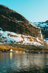 Man surfing on the surfboard on the calm sea. Snowy mountains wooden scandinavian houses and boats in the background. Summer time on the arctic.