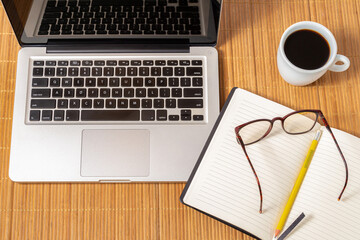 Mesa de trabalho de madeira com laptop,  um caderno de anotações aberto, com mais um óculos e uma xícara de café 