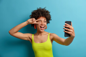 Playful positive curly haired woman makes peace sign over eye, takes selfie on smartphone, shows...