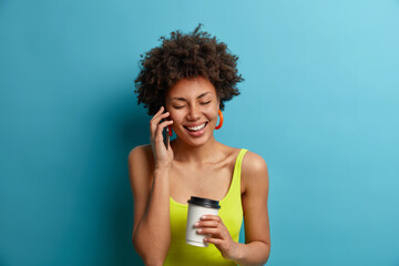 Carefree cheerful dark skinned woman with curly hair has funny amusing conversation, holds smartphone near ear, drinks takeout coffee, poses against blue background. Happy talk, technology concept