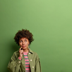 Vertical shot of curly haired young woman with thoughtful expression, keeps finger near lips, dressed in fashionable jacket, poses against green background, copy space for your advertisement