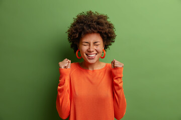 Portrait of overjoyed woman with curly hair, clenches fists with excitement, rejoices victory or...