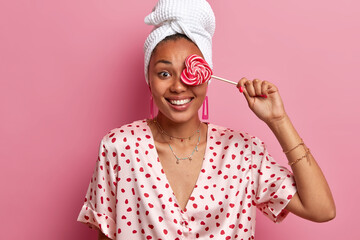 Positive ethnic dark skinned woman covers eye with sweet lollipop, wears pajama and wrapped bath...
