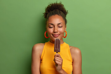 Pleased attractive woman poses for vacation photo shoot, holds delicious chocolate ice cream, closes eyes and imagines pleasant flavor, has happy time wears yellow t shirt isolated on green background - Powered by Adobe