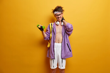 Fitness man feels pain in neck as lifts heavy dumbbell, becomes stronger every day exercises in gym dressed in sportswear, practices weight lifting poses with sport equipment against yellow background