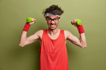 Funny sportsman puts all efforts in lifting heavy dumbbells, clenches teeth with force, does home exercises, enjoys bodybuilding, stands motivated against green background. Healthy lifestyle concept - Powered by Adobe