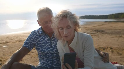 Active senior couple reviewing photos on mobile phone at beach - Powered by Adobe