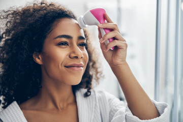 Dreamy woman applying an electric facial cleansing brush