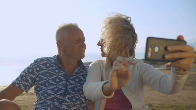 Senior couple taking selfie with mobile phone relaxing on sand at beach