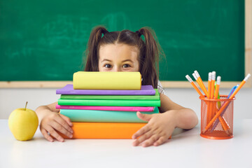 Funny little schoolgirl holds a book over her head while sitting in a lesson in class in school.