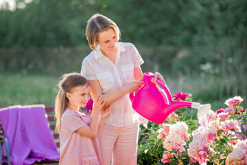 Obraz premium Mom and daughter in a linen pink dress take care of peonies in the garden at the cottage, water the flowers. He perfectly smiles and laughs. Illuminated by the rays of the evening setting sun.