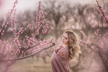 Tender fresh young girl with beautiful curly blonde hair, with a clean face, natural makeup, stands in blooming gardens, holds a twig with pink inflorescences. Mysterious, pensive, sensitive woman
