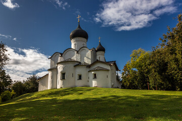 Fototapeta na wymiar Ancient white orthodox christian stone temple. Pskov region, Russia. Church of St. Basil the Great on a hill against a background of green grass and a beautiful blue sky with white clouds. 