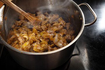 Cooking steaming stew, goulash or ragout with beef and onions in a steel pot on a black stove,...