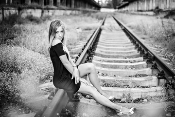 A sad and lonely girl with long hair blonde in a black short dress with long legs sits on the rails of railway tracks. The pain of loss, loss, broken heart, disappointment after parting with loved one