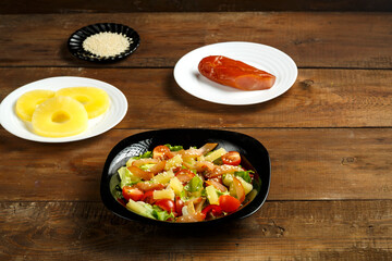Pan-Asian salad with cherry tomatoes, smoked chicken, pineapple and sesame seeds in a black plate and a range of ingredients on a wooden table.