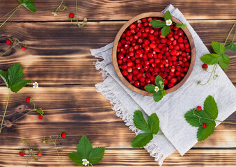 A wooden bowl of red ripe wild strawberries and flowers on old wooden surface. Top view. Copy space.
