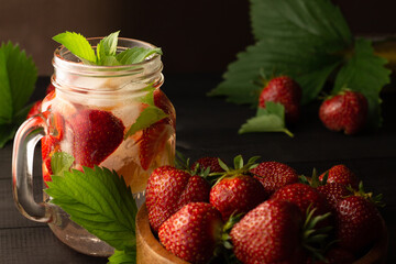 Summer cold drink with mint, ice, lemon and strawberries and wooden bowl of fresh strawberries on black wooden surface.
