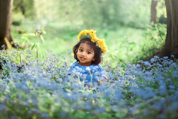 A small girl with dandelion wreath is sitting on field of forget me not blue flowers - 362235463