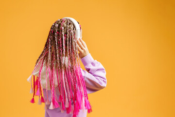 Back view of Adorable child in rounded glasses with pink dreadlocks listen to music playing in earphones on yellow background. Cute small kid listening to music in headset, copy space.