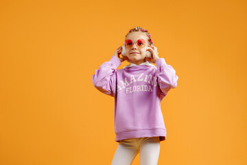 Adorable child in rounded glasses with pink dreadlocks listen to music playing in earphones on yellow background. Cute small kid listening to music in headset, copy space.