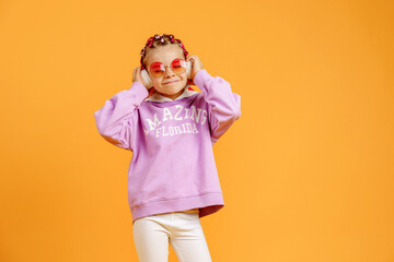 Adorable child in rounded glasses with pink dreadlocks listen to music playing in earphones on yellow background. Cute small kid listening to music in headset, copy space.