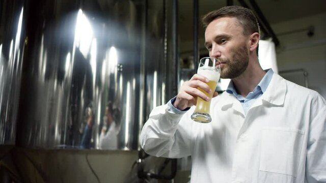 A dark-haired mature labouring man is sampling an ale that was produced at the factory. A side view slow motion close up