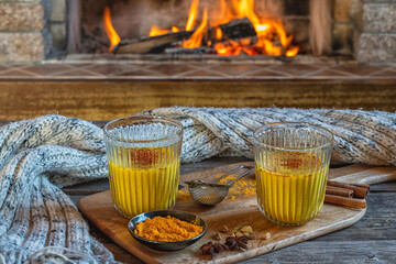 Golden latte milk in two glasses with turmeric and spices before cozy fireplace.