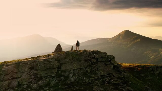 4K drone shot of climber celebrating climbing top of a mountain by raising his arms to the sky with panorama sunset background. Person standing on rock with epic mountain viewpoint