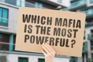 The question " Which Mafia is the most powerful? " on a banner in men's hand with blurred background. Business. Shadow government. Deaths. Illegal trade. Unlawful. Money. Control. Terror