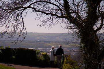 Well dressed elderly couple is enjoying the panoramic view of Bristol and the countryside,  as seen from the hill by the suspension bridge. There is an old tree near by.