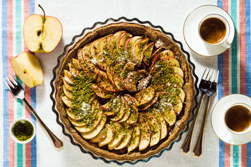 italian apple pie crostata with pistachio crumbs on the table with a white tablecloth and cups of...
