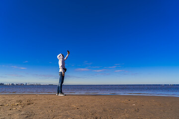 A young woman stands on the beach and films herself on a mobile phone from afar. Space for text.