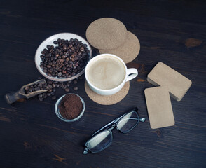 Fototapeta na wymiar Coffee cup, roasted coffee beans, ground powder, beer coasters, blank kraft business cards and glasses on kitchen table background.