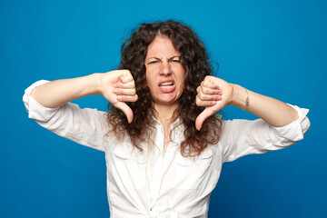 Curly haired woman shows disapproval sign, gives thumb down gesture, dislikes something, has disgusting expression, isolated over blue background. Negative human expression and body language concept.