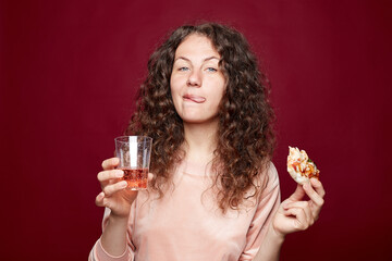 Mmm, so delicious! Curly haired pretty woman eats slice of Italian pizza, keeps tongue out from pleasure, enjoys nice taste, holding glass with bubbles, isolated on crimson background. Eating concept.