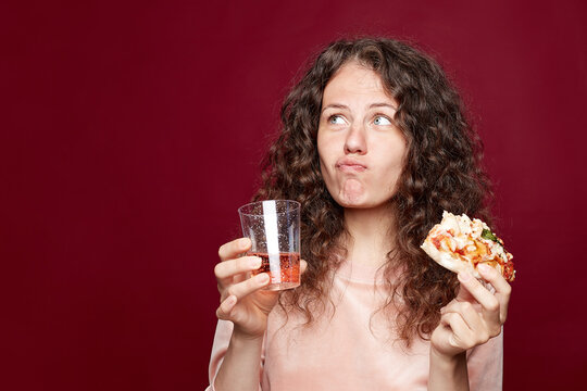 Candid close up portrait of blue eyed young female, chewing delicious pizza, being hungry, drinking pink lemonade, looking aside, posing indoors on crimson studio wall with copy space on left.