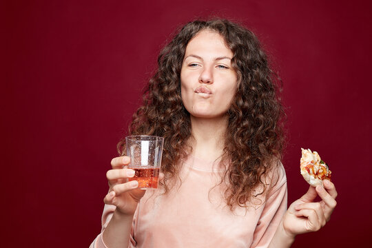 Relaxed Caucasian woman keeps slice of pizza in mouth, holds glass with bubbles, has joyful facial expression, dressed in pink sweatshirt, eats junk food, models against crimson studio background.