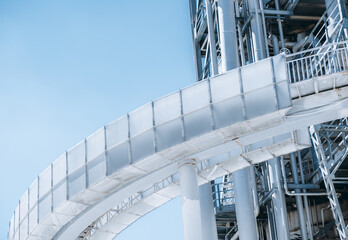 A close-up view of a contemporary construction of an industrial oil refinery or a modern fuel factory facility with a round bridge in front, multiple pipes, iron beams, and stairs, blue sky behind