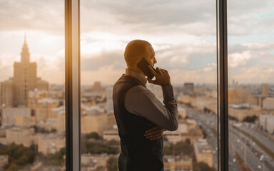 Silhouette of a hispanic businessman talking on the phone while standing near a panoramic window of a luxury office skyscraper with a roadway and a high-rise with a spire outside, beautiful sunset © skyNext