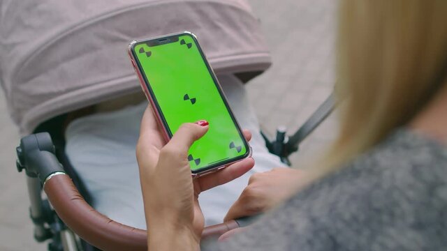 A young mother is rocking a baby stroller with one hand and flipping through a green-screen phone with the other. A girl flips through a smartphone with a chromakey on the background of a stroller