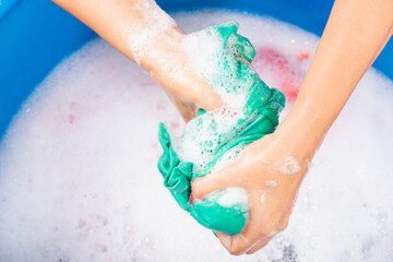 Closeup young Asian woman use hands washing color clothes in basin. Female squeeze wring out wet fabric cloth with detergent have soapy bubble in water, studio shot background, laundry concept