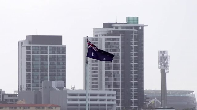 The Australian flag flies in high winds with close up of Perth skyscrapers providing the backdrop.  Optus stadium and lights of WACA ground partly visible in background