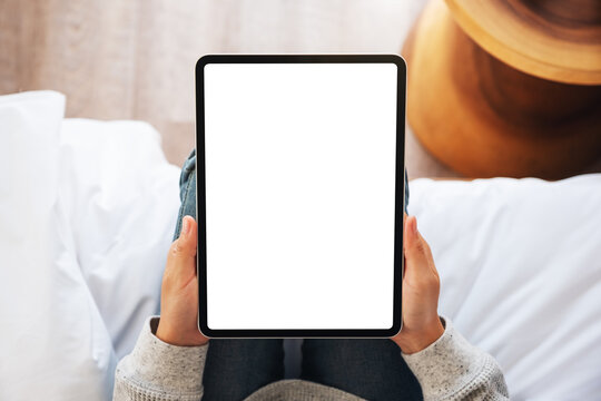 Top view mockup image of a woman holding black tablet pc with blank desktop white screen while sitting on a cozy white bed at home - Powered by Adobe