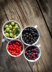 Summer berries (gooseberry red currant, black currant, shadberries) in white bowls on the old rustic wooden table, top view. 