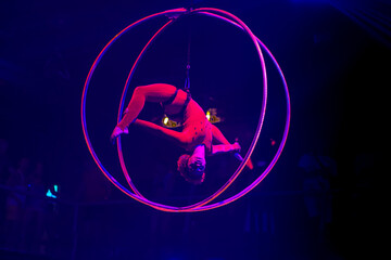 Flexible young woman make performance on aerial hoop, flexible back on aerial hoop, aerial circus show, blue and red light. Flexible woman gymnast upside down on hoop. Night club performance