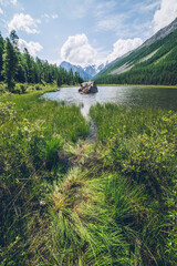 Meditative view to beautiful lake with stone in valley on snowy mountains background. Scenic relaxing green landscape with big mossy stone in mountain lake. Alpine lake with ripples on water surface.
