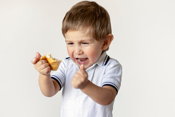 A 2-year-old boy opened his mouth, shouts, shakes his hand, in which he holds a pie. White background. Close-up